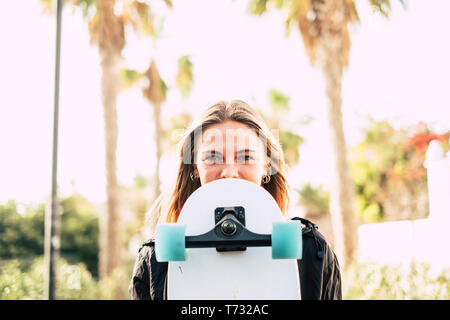 Modern cheerful beautiful blonde girl portrait with skateboard covering part of the pretty face - young millennial people outdoor concept enjoying lif Stock Photo