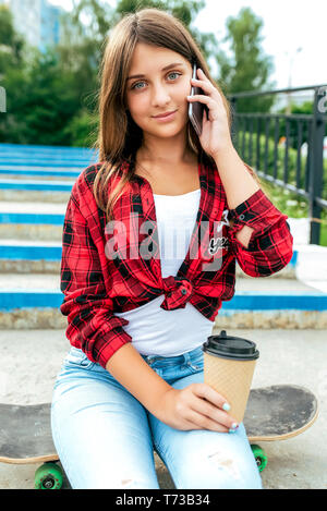 Girl teenager sits on a skateboard, talking on the phone, holding a mug with tea in her hands. In the summer in the city in nature. Stock Photo
