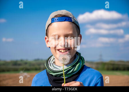 boy with shield cap blows a flowering dandelion in front of a blue sky Stock Photo