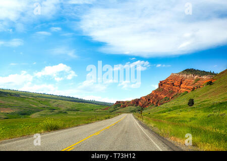 Tiered red rock formation jutting toward a winding highway between rolling hills along Chief Joseph scenic highway in a summer time Montana landscape Stock Photo