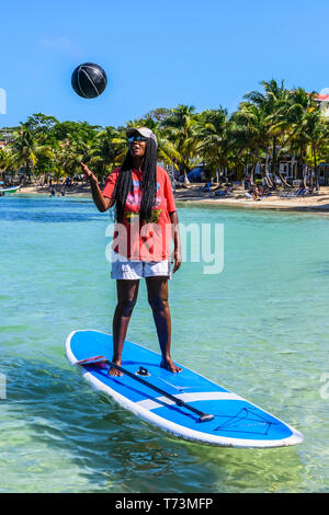 A woman stands up a paddle board tossing a ball in the air off the coast of West End Village; Roatan, Bay Islands Department, Honduras Stock Photo