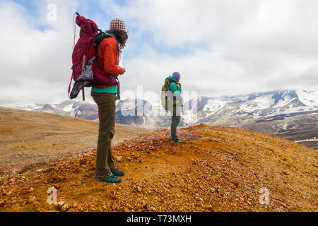 Two women backpacking and taking in the view of Mount Katmai from the rainbow-coloured pumice, rock and clay slopes of Broken Mountain, Valley of T... Stock Photo
