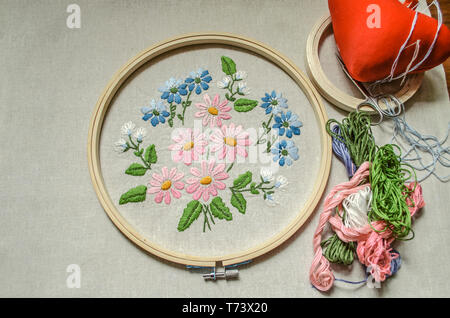 Cotton fabric with embroidered bouquet of wildflowers covered with wooden hoops, colored threads, scissors and a red pillow for needles Stock Photo