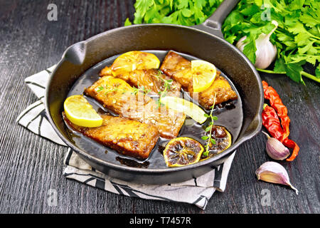 Pink salmon with honey, lemon juice, garlic, hot pepper and soy sauce, lemon slices and a sprig of thyme in frying pan on a napkin, parsley on black w Stock Photo