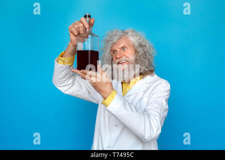 Old long-haired man wearing doctor's gown studio isolated on blue wall standing holding bottle of spirit looking at drink passionate Stock Photo