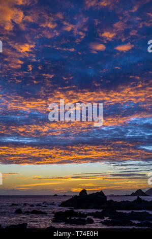 Glowing, golden clouds and silhouettes of rock formation and rugged coastline along the South coast at sunset; Wellington, New Zealand Stock Photo