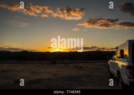The sun is setting behind a range of hills, lighting up the few clouds in orange and red hues. The side of a camper truck is visible on the right h... Stock Photo
