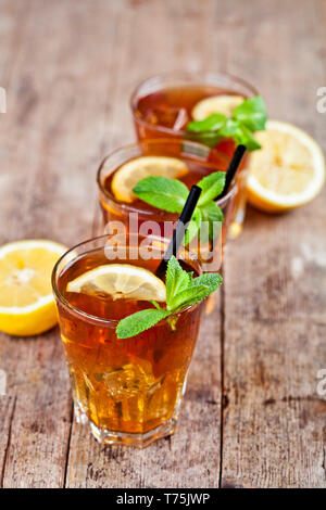 Cold iced tea with lemon, mint leaves and ice cubes in three glasses on rustic wooden table background. Stock Photo