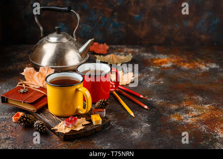 Two colored red and yellow enamel cups of tea, watercolors in cuvettes, colored pencils, autumn maple leaves and bumps on a rusty brown background Stock Photo