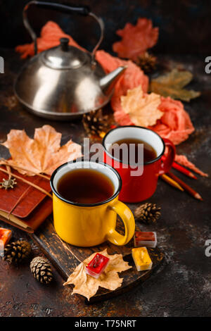 Two colored red and yellow enamel cups of tea, watercolors in cuvettes, colored pencils, autumn maple leaves and bumps on a rusty brown background Stock Photo