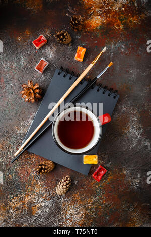 Red enamel cups of tea, watercolors in cuvettes, brushes and bumps on a rusty brown background Stock Photo
