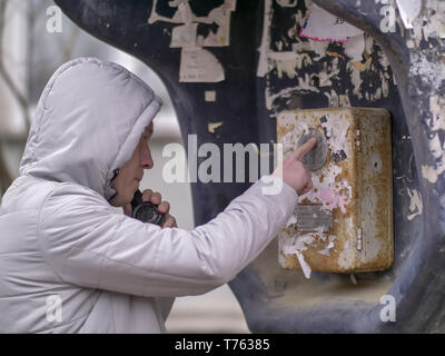 Man in a gray jacket with a hood is calling from an old payphone on the street Stock Photo