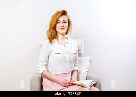 Young beautiful caucasian woman with long magnificent red hair and freckles on face. Student girl sitting on armchair and holding book in hands, a man Stock Photo