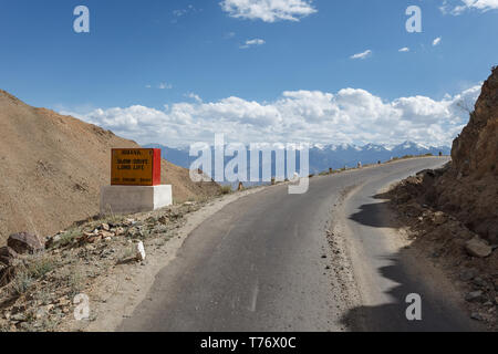 Paved section of road high in snow capped mountains rounding curve on pass through mountains in Kashmir, India on a sunny day with sign reminding peop Stock Photo