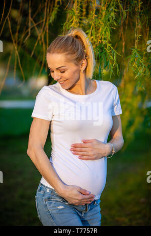 Portrait of a happy and proud pregnant young woman looking at her belly in park at sunrise, new life concept Stock Photo