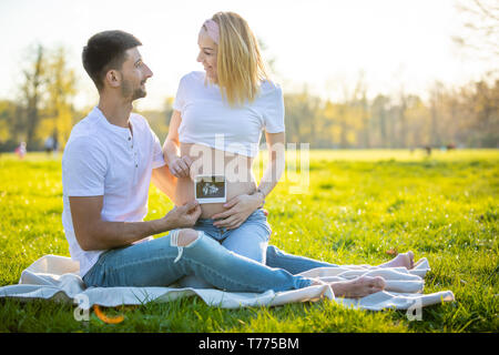 Happy couple expecting baby, pregnant young woman with husband sitting on grass and holding ultrasound pictures, young family and new life concept Stock Photo