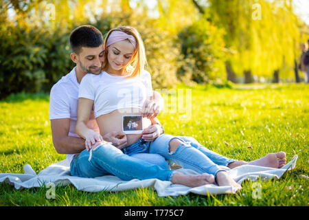 Happy couple expecting baby, pregnant young woman with husband sitting on grass and holding ultrasound pictures, young family and new life concept Stock Photo