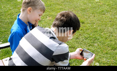 teen boys with smart phone listening or talking in british park on bench. teenager and social media concept. Stock Photo