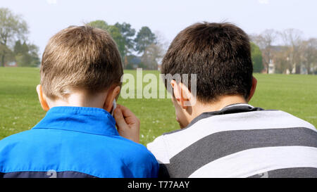 teen boys with smart phone listening or talking in british park on bench. teenager and social media concept. Stock Photo