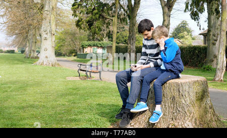 teen boys with smart phone listening or talking in british park on bench. teenager and social media concept. Stock Photo