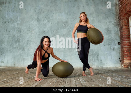 Two young, beautiful fitness girls in the gym posing with fitness balls against a gray wall Stock Photo