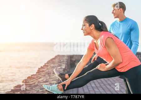 Sporty couple stretching legs in front of ocean at sunset - young people training and making legs exercises outdoor Stock Photo