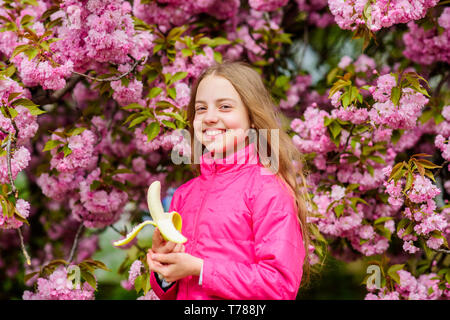 Little girl eat banana. Kid on pink flowers of sakura tree background. Kid enjoying cherry blossom sakura. Happy spring vacation. Spring in botany garden. That is how spring smells. Tender bloom. Stock Photo