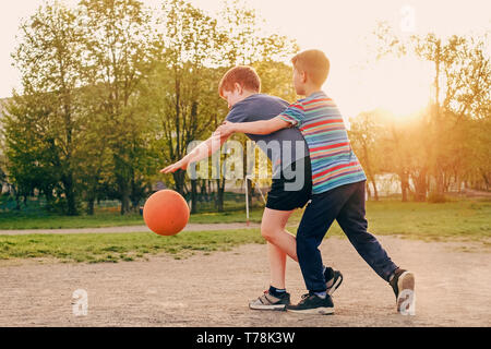 Two happy young boys playing basketball outdoors on a sports field in spring backlit by the warm glow of the evening sun Stock Photo