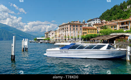 A tourist boat moored in Bellagio harbour on Lake Como, with the Hotel Metropole and other colourful buildings beyond under a bright blue sky Stock Photo