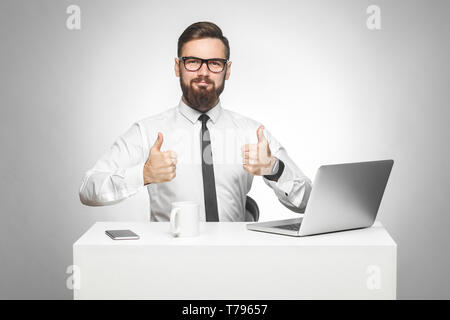 Everything alright! Portrait of handsome satisfied bearded young businessman in white shirt and black tie are sitting in office and working on laptop  Stock Photo