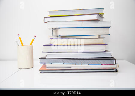 Home office desk with a high stack of colourful books in a pile and a container with pens and pencils. White desk gives a light, bright airy feeling. Stock Photo