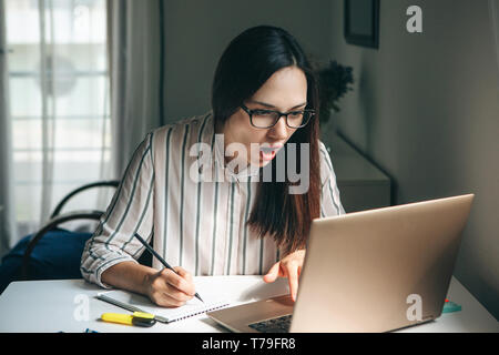 A young beautiful student girl does homework or works in a home office. She is surprised by the news or she has found an unexpected solution. Stock Photo