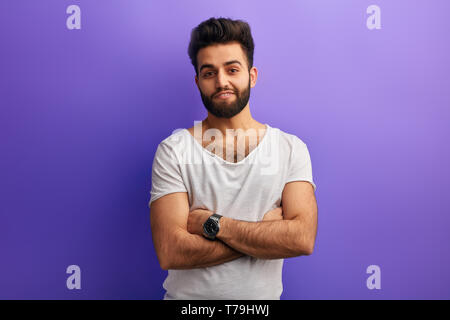 bearded Indian man stands with crossed arms, expresses positive emotion against blue background. people, free time, spare time. guy in white T-shirt w Stock Photo