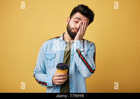 Tired exhausted student with sleepy expression, covers face with palm, holds disposable paper cup of coffee. man doesn't want to go to work. isolated  Stock Photo