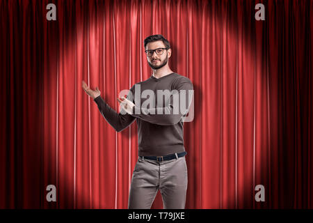 Front crop view of young handsome man in casual clothes and glasses standing in spotlight against red stage curtain and making presenting gesture at Stock Photo