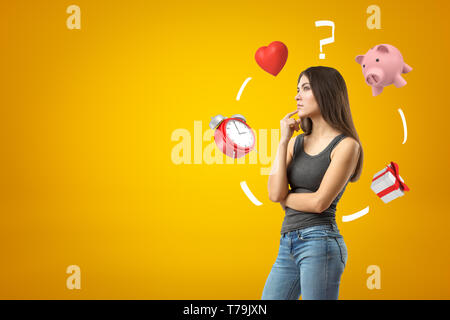 Young brunette girl wearing casual jeans and t-shirt thinkings with alarm clock, red heart, question mark, piggy bank and gift box on yellow Stock Photo