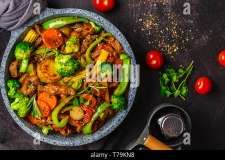 Fried beef stroganoff with potatoes, broccoli, corn, pepper, carrots and sauce in a pan, dark background. Stock Photo