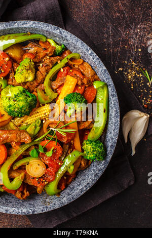 Fried beef stroganoff with potatoes, broccoli, corn, pepper, carrots and sauce in a pan, dark background.