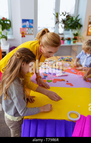 Girl near teacher. Cute dark-haired girl standing near teacher while painting on yellow paper Stock Photo