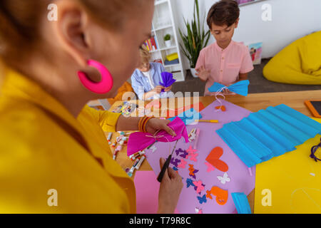 Making paper bow. Teacher using scissors while making paper bow out of pink paper while standing near pupils Stock Photo