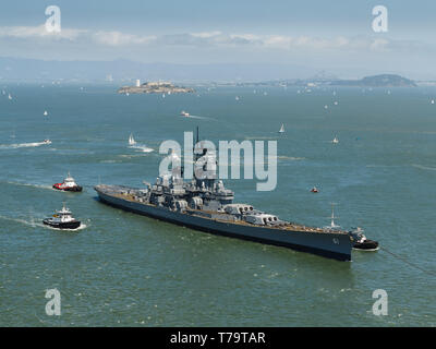 Closeup view of the side of a navy world war II battleship, and tugboats leaving San Francisco harbor with hundreds of sailboats dotting water under s Stock Photo