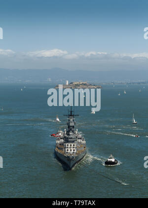 View of San Francisco harbor with hundreds of sailboats dotting water under sail, closeup front a navy world war II battleship, and tugboats leaving b Stock Photo