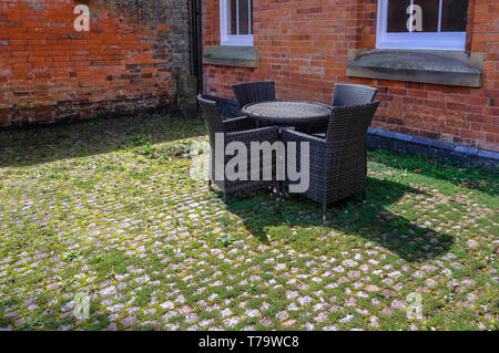 Wicker table and arm chairs  in dark grey set in an old courtyard witha red brick background.  Weeds and moss between the cobbles add to a traditioana Stock Photo