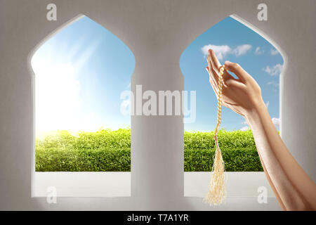 Muslim hands praying with prayer beads in the mosque with garden view from window arches over blue sky background Stock Photo