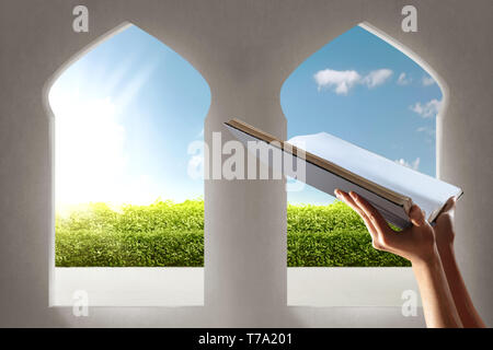 Muslim hands holding quran open in the mosque with garden view from window arches over blue sky background Stock Photo