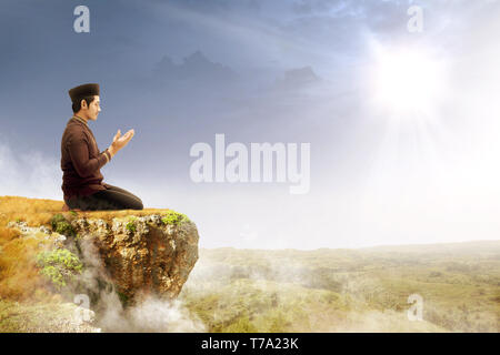 Asian muslim man sitting in pray position while raised hands and praying on the edge of the cliff with sunlight and landscapes background Stock Photo