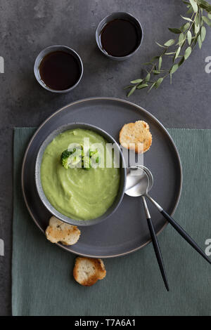 Broccoli cream soup with bread and spoon on gray plates, with tea cups and green napkin on gray background. Concept of healthy food. Flat lay, top vie Stock Photo