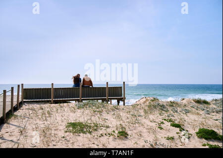 Young couple seated on a bench watching the ocean with the wind on their hair, over the dunes Stock Photo