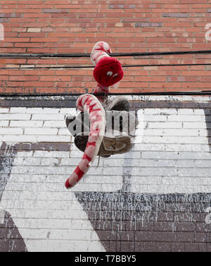 Dangling tennis shoes and a stuffed toy animal snake hanging from a power line with a painted brick wall in the background. Stock Photo