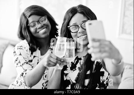 Two african woman friends wear on eyeglasses posed indoor white room, drinking champagne and making selfie. Stock Photo